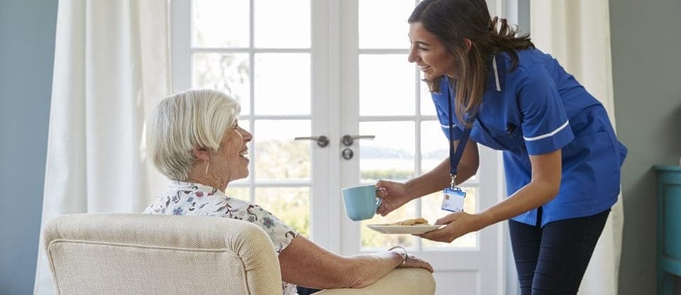 A nurse bringing a cup of tea to a resident seated by a sunny window